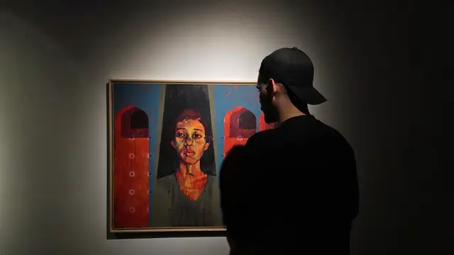 Amir in profile, wearing a black cap and dark clothes, standing in a dim gallery and looking at a framed portrait painting of a woman against orange arches.