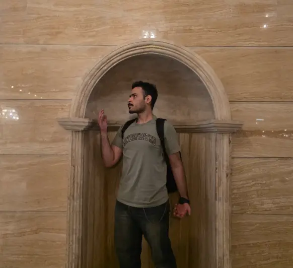 Amir standing in a marble archway, in a grey T-shirt with a backpack, looking off to the side.